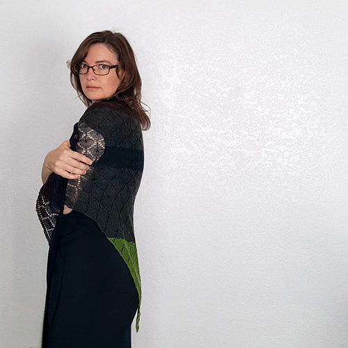 Side-back view of model looking over her shoulder wearing the Kellerwald shawl in green and black, showing the black lace section across her back with green edge visible below
