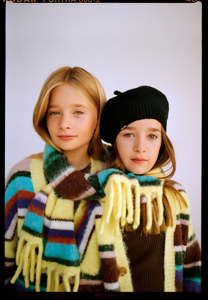 Two children wearing colorful scarves and hats against a plain background