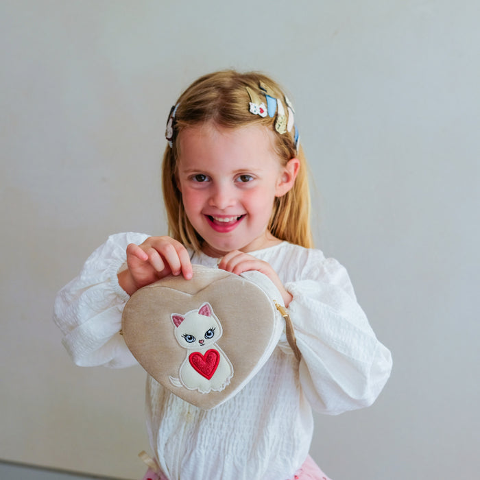 A young girl with blonde hair in a white ruffled blouse holds a heart-shaped beige cushion featuring a white cat with a red heart on the front, set against a plain light gray wall.