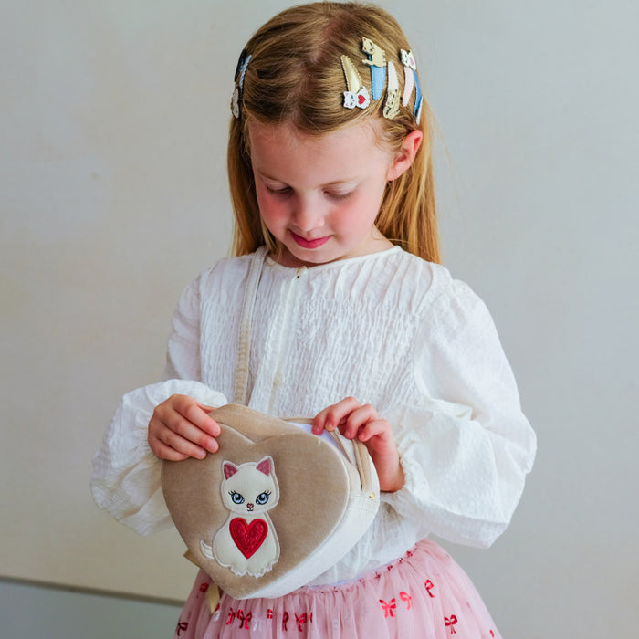 A young girl with blonde hair in a white smocked blouse and pink skirt holds a beige heart-shaped fabric bag with a white cat and red heart applique, set against a plain light gray background.