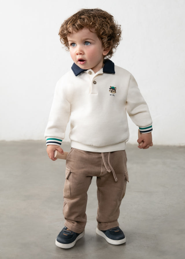 Child wearing a white sweater with a logo and brown pants on a plain background