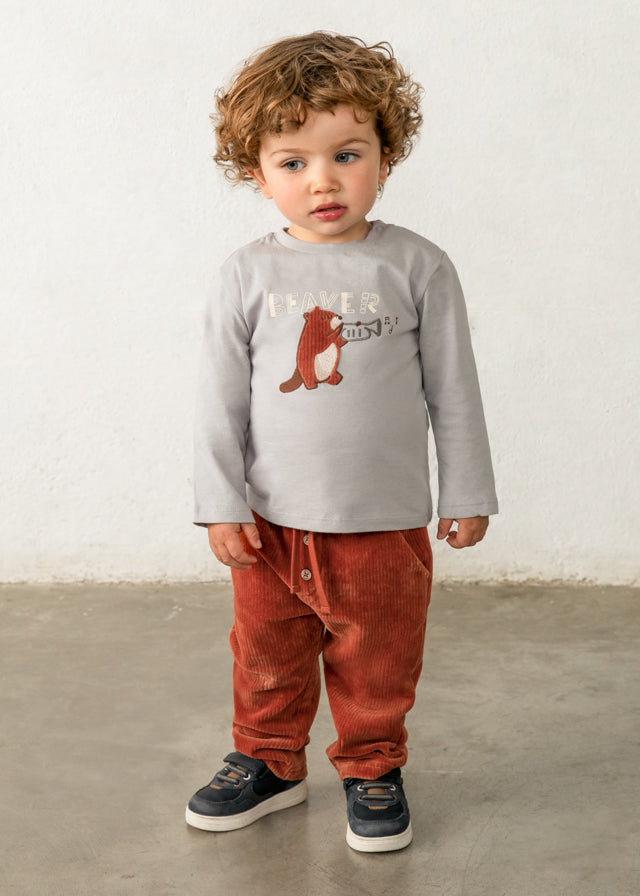 Child wearing a gray long-sleeve shirt with a bear graphic and red corduroy pants against a white wall.
