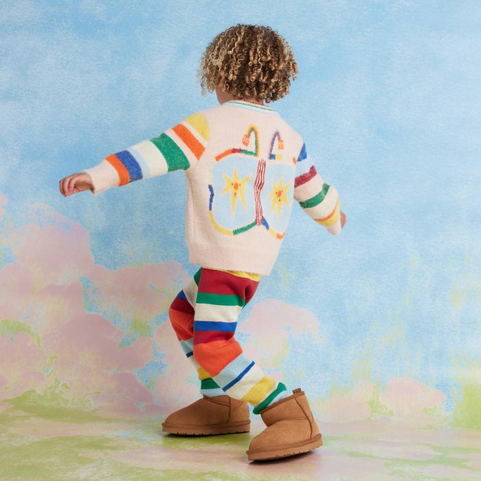 Child wearing a colorful outfit with abstract designs, standing on a grassy field with a sky background.