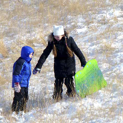 Girl in a white knit hat and black coat holding hands with a young child in blue winter gear while walking up a snowy hillside carrying a green sled, both wearing hand-knit striped mittens