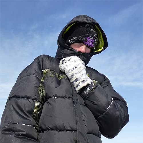 Person bundled in a black puffer jacket and hood in the snow, holding up a white and gray striped hand-knit Family Mitten near their face against a blue winter sky