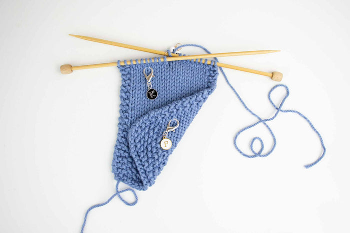 Wider view of knit and purl reminder markers on blue knitting swatch with bamboo needles and yarn trailing