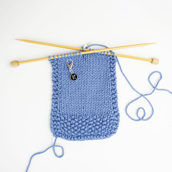 K reminder clip marker attached to stockinette side of blue knitting on bamboo needle