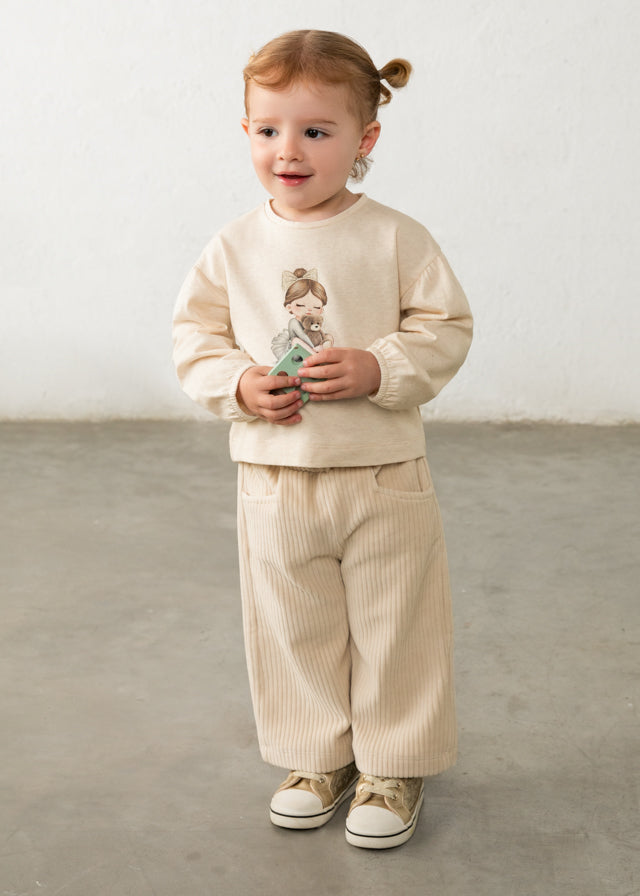 Child wearing a beige outfit with a graphic design, standing on a plain floor.