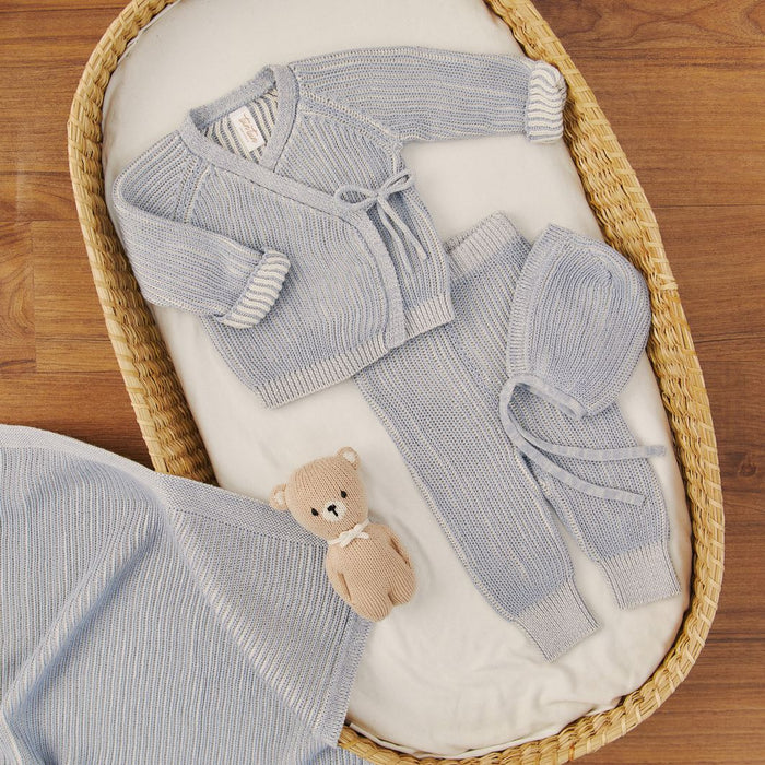 Knitted baby outfit with hat and bear toy in a wicker basket on a wooden floor.