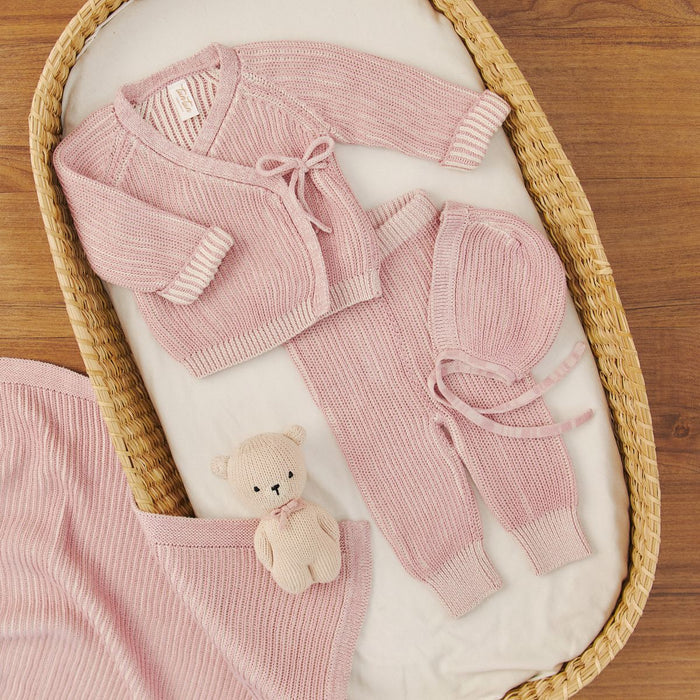 Pink baby outfit with matching blanket and teddy bear in a wicker basket on a wooden floor.