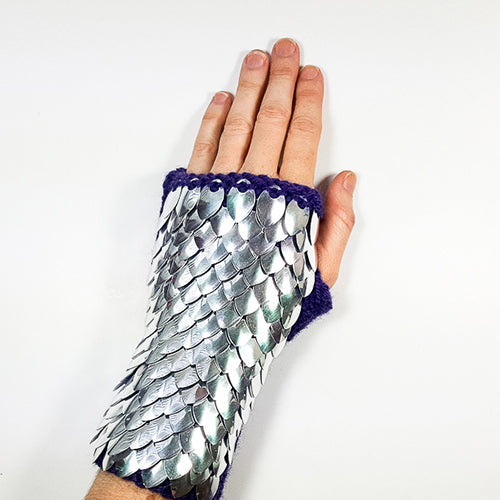 Hand wearing a silver metallic dragonscale fingerless gauntlet with purple knitted cuff, shown palm-down on white background
