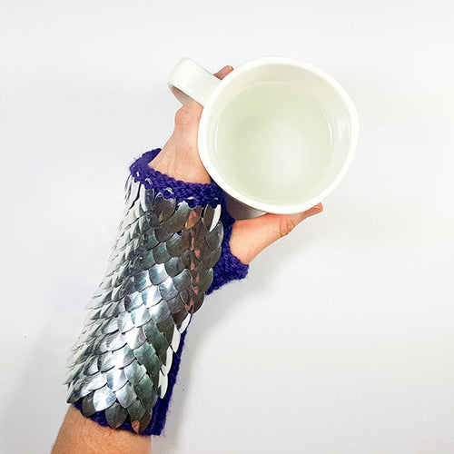 Hand wearing silver metallic dragonscale fingerless gauntlet with purple knitted cuff, holding a white coffee mug to show the underside and flexibility