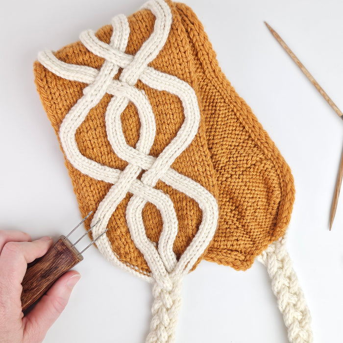 Hand holding a golden knit swatch with cream freeform i-cord cable designs applied using the Cord Quill, showing the decorative raised cable technique