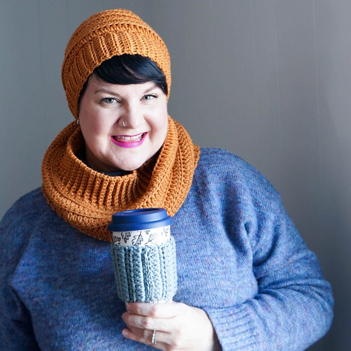 Woman wearing golden mustard crochet cowl and matching hat with sage cup cozy, holding travel mug