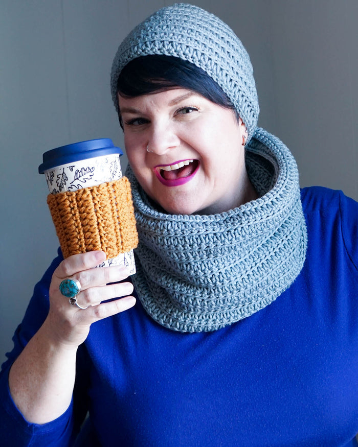 Woman wearing dusty blue crochet hat, cowl, and mustard cup cozy, smiling with travel mug