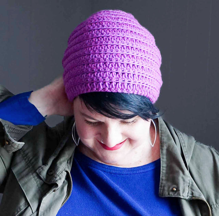 Woman wearing purple textured crochet beanie with blue shirt and olive jacket against gray background