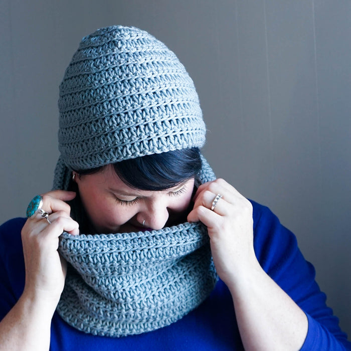 Woman wearing dusty blue crochet hat and matching cowl set, snuggling into cowl against gray background