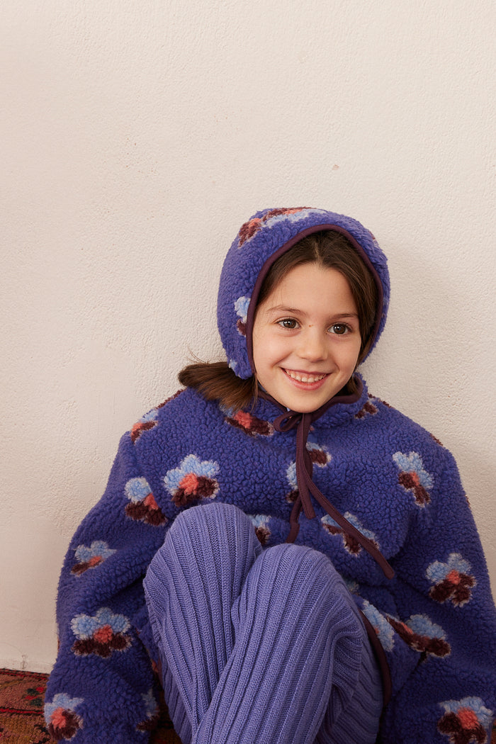 A young girl smiling while wearing a cozy, fuzzy purple hat and matching jacket with ribbed purple pants against a plain off-white wall.