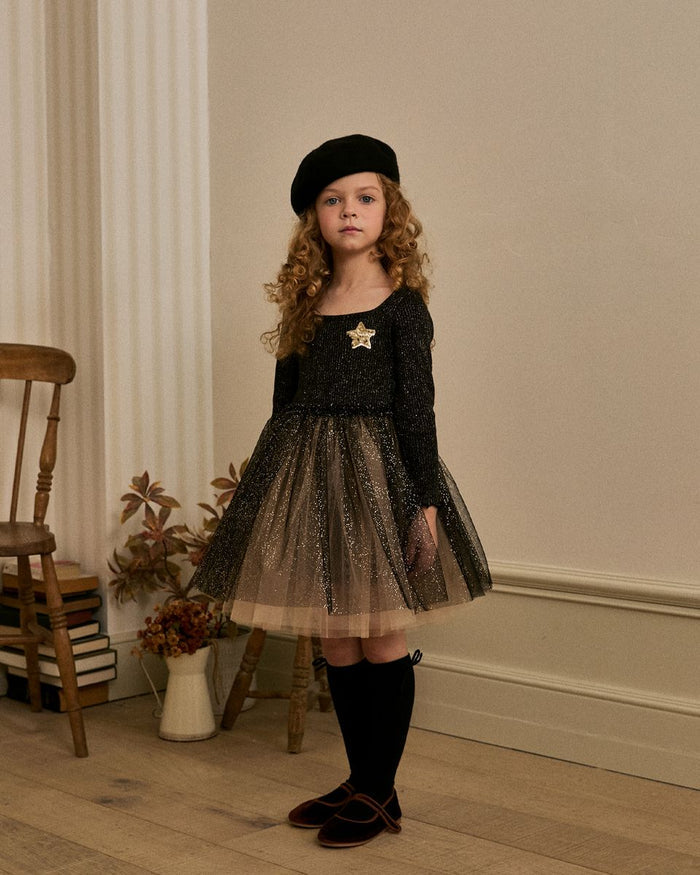 Young girl in a black dress with a star and a beret standing in a room with wooden floor and chair.