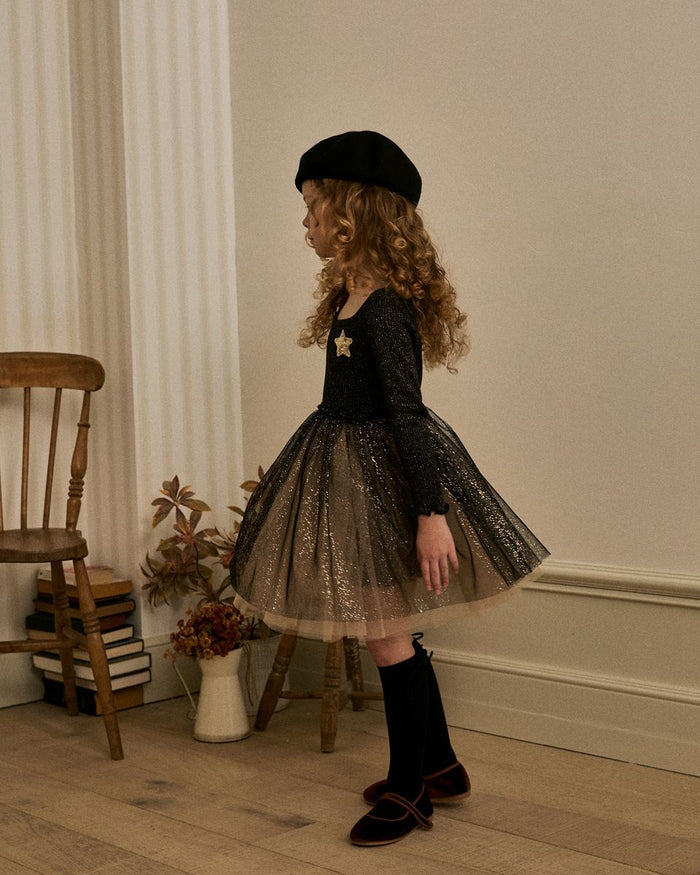 Young girl in a black dress with a tutu skirt and beret standing in a room with wooden floor and white walls.