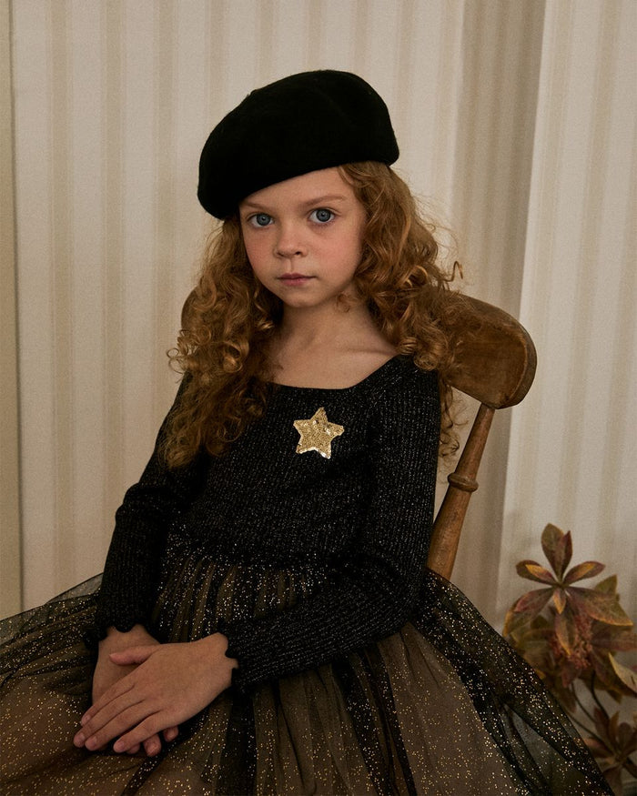Young girl in a black dress with a gold star and black beret sitting on a wooden chair.