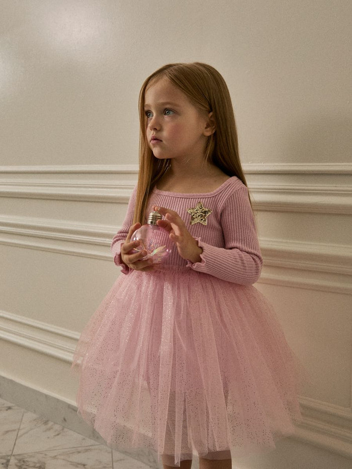 Young girl in a pink dress holding a clear jar with a star decoration against a neutral wall.