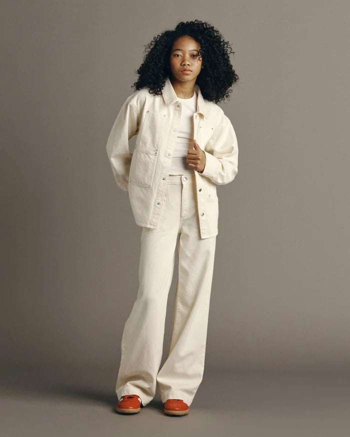 A model with curly black hair stands against a solid gray studio background wearing a matching off-white denim jacket and wide-leg pants set, styled with a white ribbed t-shirt and orange slip-on shoes.