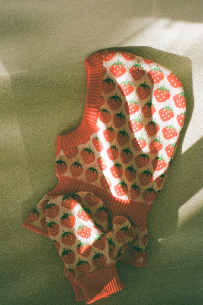 A knit balaclava and mittens with strawberry patterns , laid flat on a textured olive-green surface with dappled sunlight and shadows.