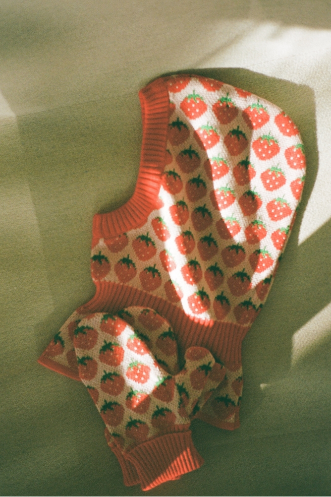 knit balaclava and mittens with strawberry patterns, laid flat on a textured olive-green surface with dappled sunlight and shadows.