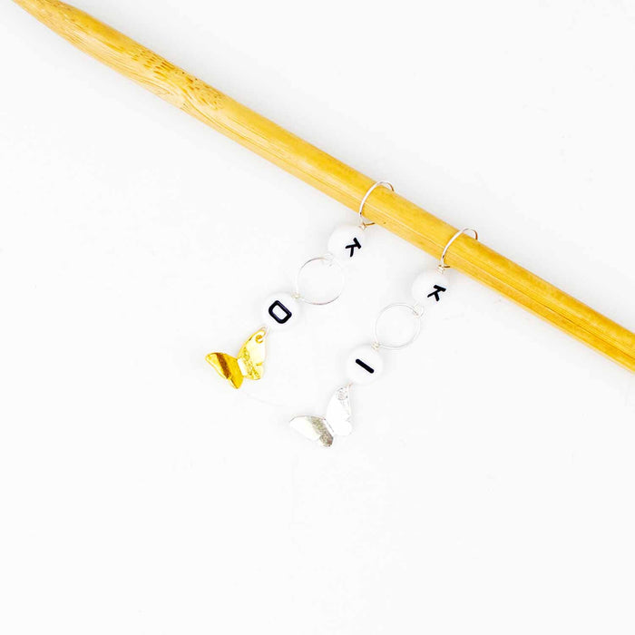 Little butterflies increase/decrease marker set on blue knit fabric, showing colorful enamel butterfly charms with K, I, and D letter bead markers on the knitting needle