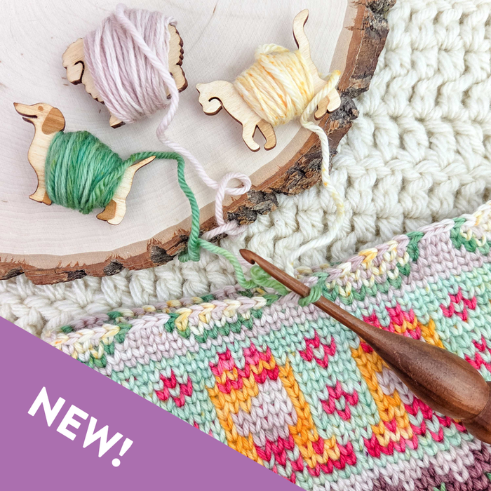 Three wooden animal yarn bobbins wrapped with green, lavender, and yellow yarn on a wood slice with yarn tails feeding into a colorwork crochet project with floral motif and a wooden crochet hook