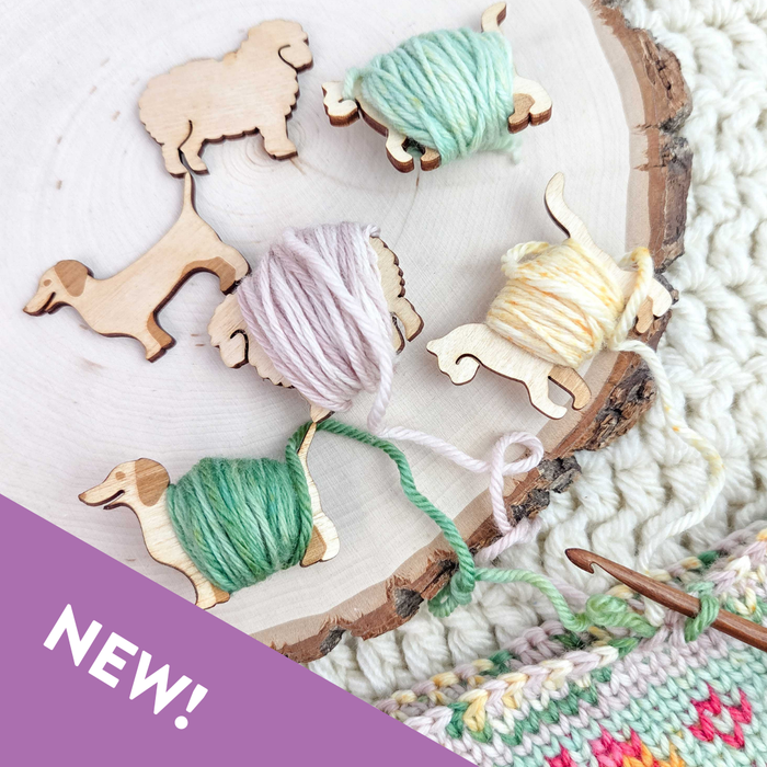 Five wooden animal yarn bobbins including cats, dogs, and sheep on a natural wood slice, some wrapped with green, lavender, and yellow yarn, alongside a colorwork crochet piece with one large unwrapped sheep bobbin