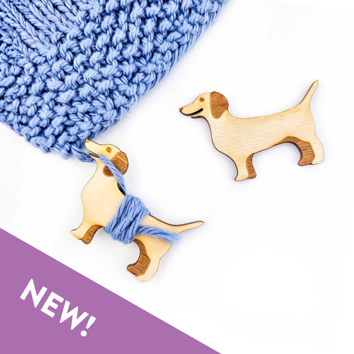Two laser-cut wooden dachshund-shaped yarn bobbins next to blue knitted fabric on a white background with one bobbin holding blue yarn wrapped around its body and the other empty showing natural birch wood grain