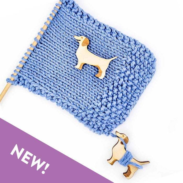 Blue knitted swatch on a knitting needle with a wooden dachshund yarn bobbin resting on top of the fabric and a second bobbin with blue yarn wrapped around its body dangling from the yarn tail below