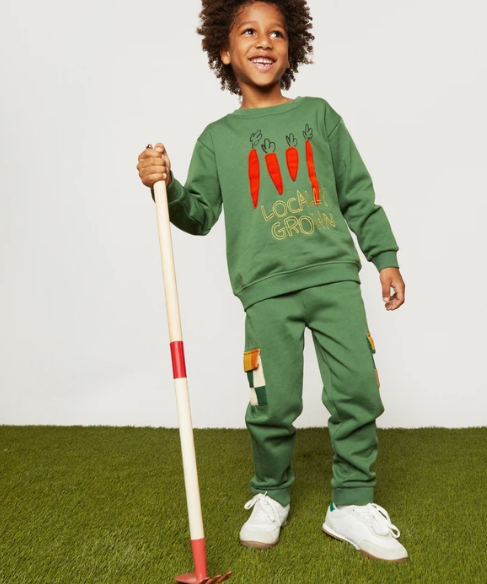 A young child wearing a green sweatshirt and matching sweatpants set, holding a garden hoe prop. The sweatshirt features red embroidered carrots and the text 'LOCAL GROWN'.