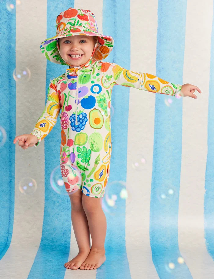 Child wearing a colorful fruit-patterned swimsuit and hat against a striped background