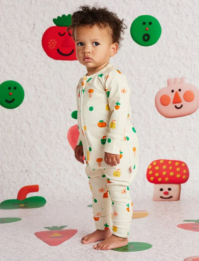 Child wearing a onesie with colorful vegetable patterns against a wall with cartoonish vegetable decorations.