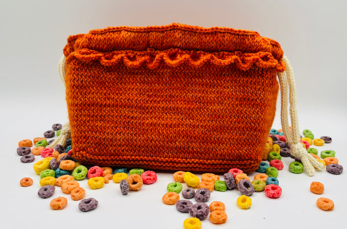 Finished Loopdedoo Dumplin Bag in bright orange yarn with drawstring closure, shown with colorful candy pieces scattered around it