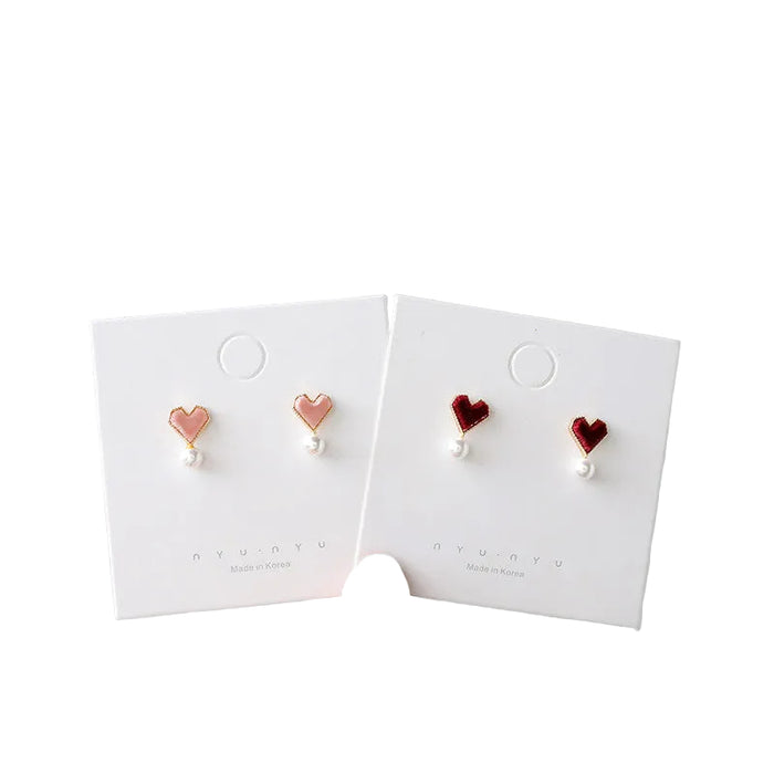 Two pairs of heart-shaped stud earrings displayed on white "nyunyu" branded cards. One pair features light pink hearts with a gold border and a small white pearl dangling below, while the other pair features deep burgundy velvet hearts with similar pearl accents. Both are labeled "Made in Korea."