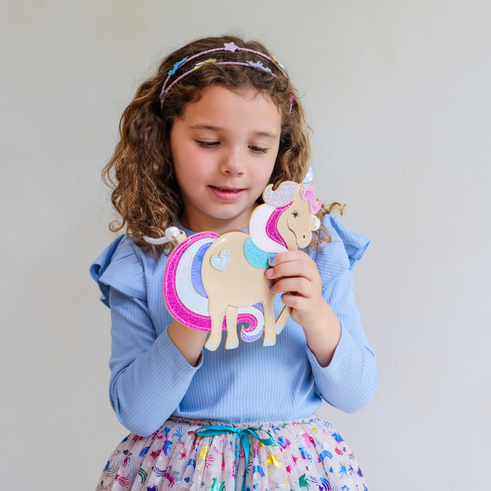 A young girl with curly brown hair wearing a light blue top and a colorful skirt, holding a unicorn purse with glittery pink, blue, and white details against a plain light gray background.