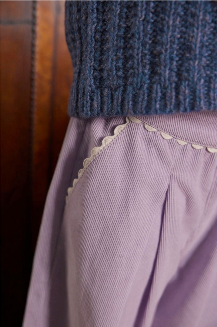 Close-up of a person wearing a dark blue ribbed knit top and a lavender corduroy skirt with cream scalloped trim, against a dark brown leather background.