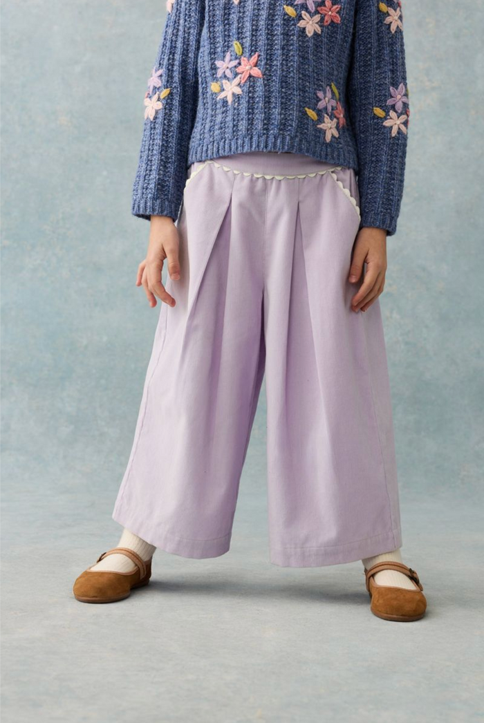 A child wearing wide-leg lavender pants with a decorative white beaded waistband, a blue floral knit sweater, and brown flat shoes, standing against a textured light blue backdrop.