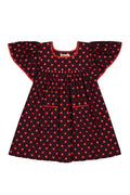A toddler girl's dress featuring a dark red base with a red-orange polka dot pattern, ruffled cap sleeves, and a smocked neckline, shown against a plain white background.