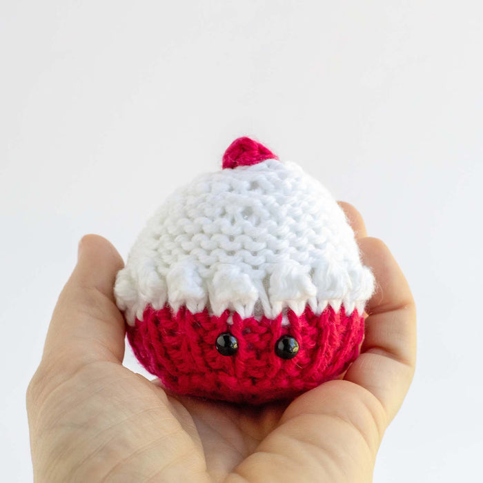 Hand holding small knitted pink and white cupcake softie with black safety eyes and cherry on top