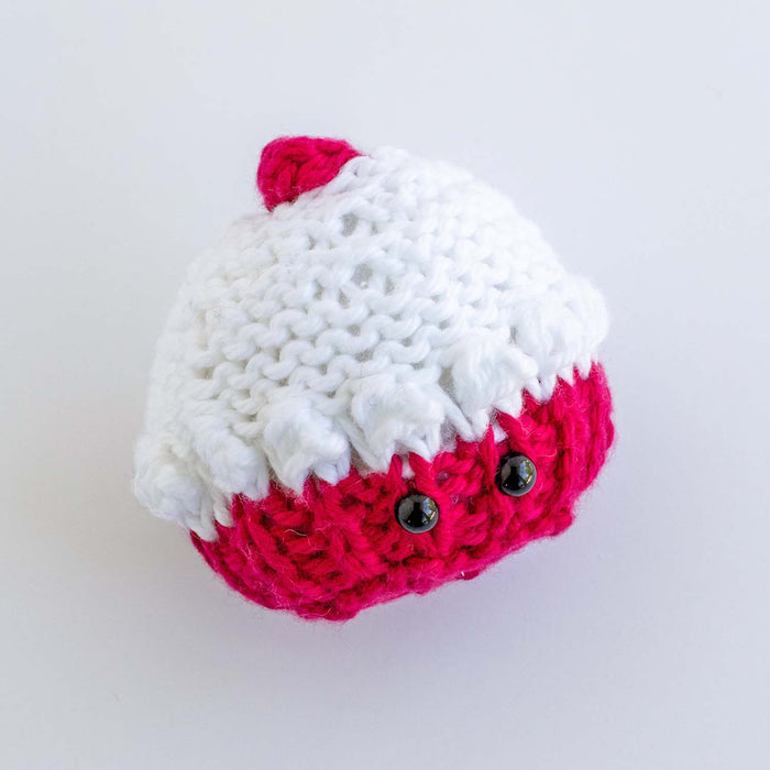 Knitted pink and white mini cupcake softie with safety eyes and cherry topper viewed from above