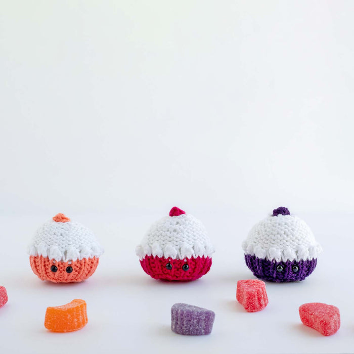 Three knitted mini cupcake softies in orange, pink, and purple with white frosting tops surrounded by gummy candies