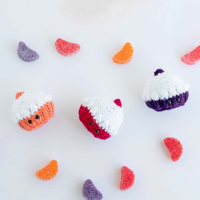 Three knitted mini cupcake softies tumbled on their sides with colorful gummy candies scattered around them