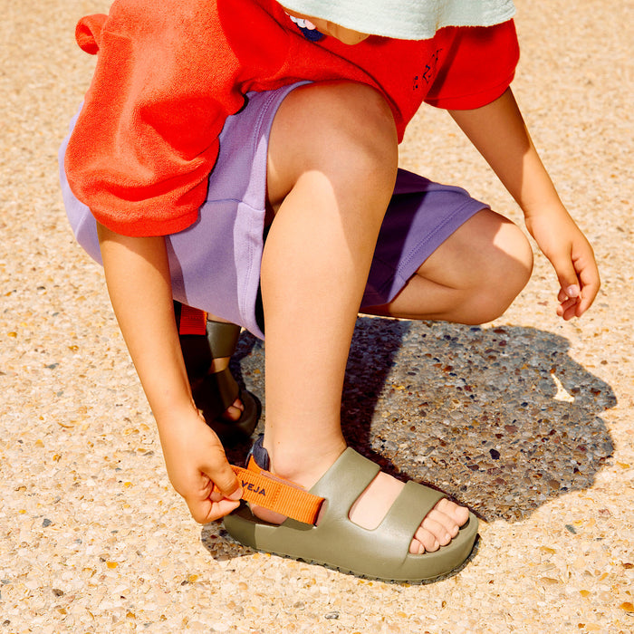 A child wearing an olive green sandal with an orange strap, crouching on a sandy ground while adjusting the strap. The brand name 'VEJA' is visible on the strap.