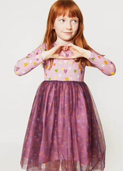 A young girl with red hair wearing a pink long-sleeve dress with a heart pattern and a sheer purple tulle skirt, posing with her hands together against a light gray background.