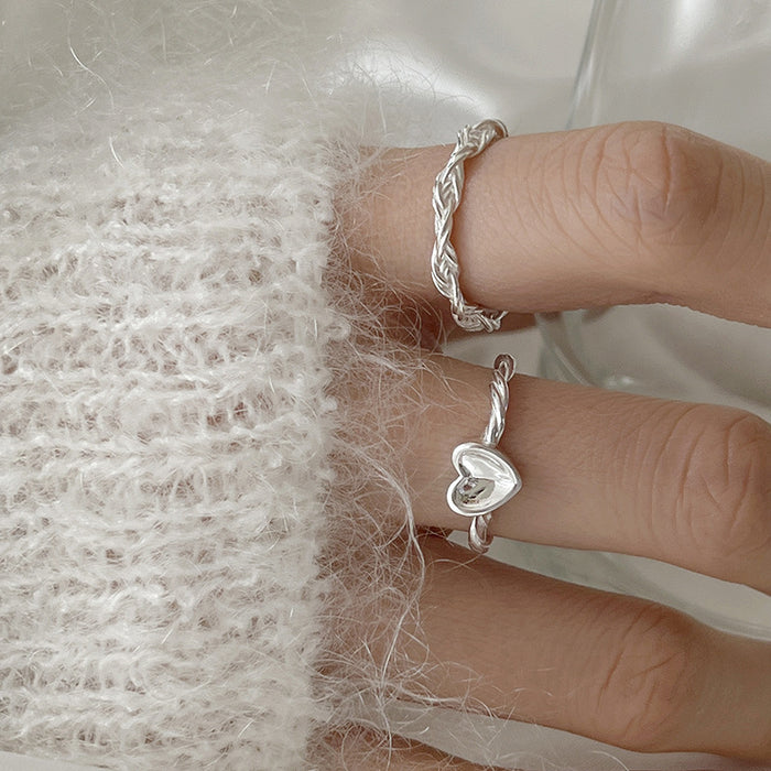 Two silver-tone rings worn on a hand against a soft white knit background; one ring features a braided rope design and the other a matching braided band with a central polished heart charm.